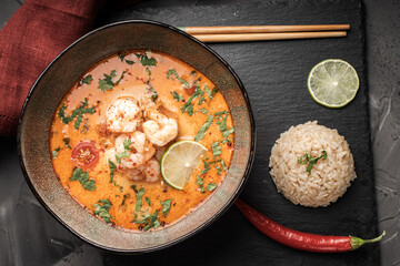 Tom Yum Kung Spicy Thai soup with shrimp in a bowl on a dark background and red pepper, rice and lime with cilantro and chopsticks on a black mica board. Copyspace