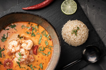 Tom Yam Kung Spicy Thai soup with shrimp in a bowl on a dark background and red pepper, rice and lime with cilantro and a spoon on a black mica board.