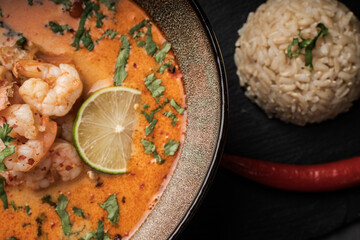 Tom Yum Kung Spicy Thai soup with shrimp and cilantro in a bowl on a dark background with rice and red hot pepper on a black mica board. Top view, close-up. Copyspace.