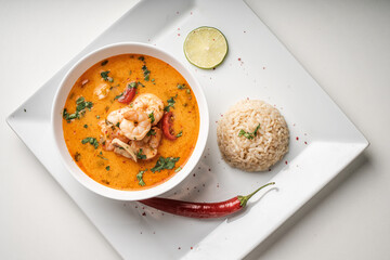 Tom Yam Kung Spicy Thai soup with shrimp and cilantro in a white bowl on a light background with rice and red hot pepper, lime and chopsticks on a square plate. Top view, close-up. Copyspace.