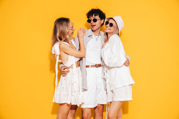 Group of young three stylish friends posing in studio. Fashion man and two cute female dressed in casual summer clothes. Smiling models having fun. Cheerful women and guy near yellow wall, sunglasses