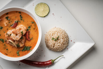 Tom Yam Kung Spicy Thai soup with shrimp and cilantro in a white bowl on a light background with rice and red hot pepper, lime on a square plate. Top view, close-up. Copyspace.