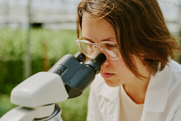 Scientist intently observing sample through microscope in laboratory setting wearing white lab coat and safety glasses, background blurred with laboratory equipment