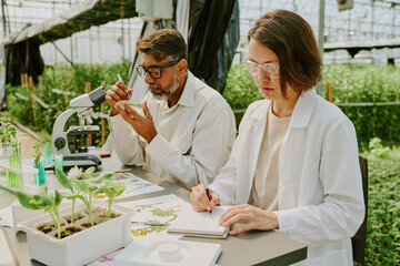 Two scientists working in a lab examining plant specimens surrounded by lush greenery. Researching...