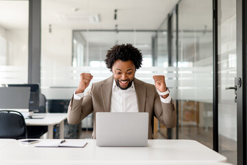 An enthusiastic African-American businessman celebrates a successful moment at his laptop, his...