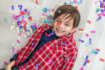Happy young boy lying on floor with colorful confetti scattered around, celebrating smiling and enjoying the festive moment, cheerful child in red plaid shirt, happiness, joy, fun, kids party, playful