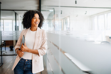 A Brazilian businesswoman smiles while looking to the side, standing in an office hallway that...