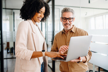 A businesswoman and her mature male colleague explore ideas on a laptop, standing in an office scene that. Image combines expertise and modern tech, blend of wisdom and current technology trends