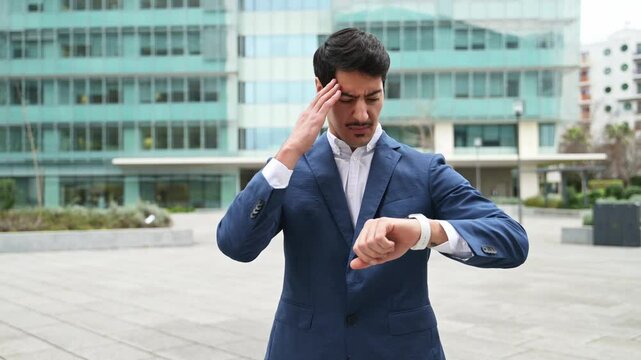 A Hispanic businessman in a blue suit looks concerned while checking the time on his watch outside a corporate building, representing the fast-paced nature of business, feels leak of time and deadline