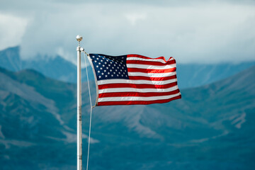 The USA American flag waving proudly with the Alaskan mountains in the background on a cloudy summer day