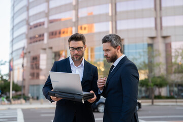 Two entrepreneurs near the office on the street. Business meeting of friends outdoors. Business mens with takeaway coffee and laptop outdoor. Business men team using laptop outdoor. Two businessman