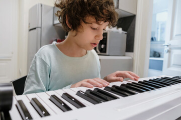 Focused young boy with curly hair playing digital piano at home in kitchen learning music skills