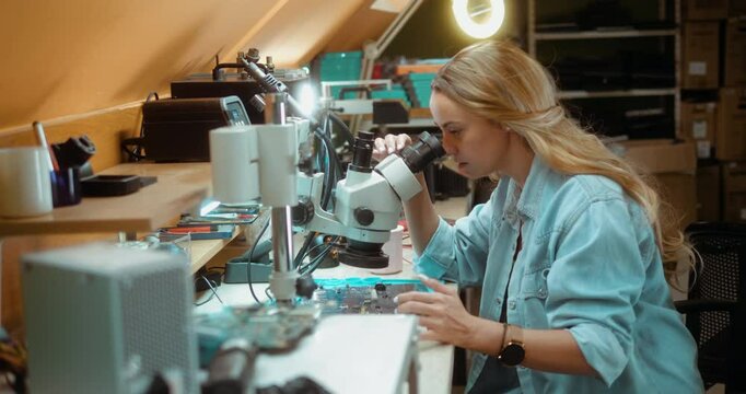Female technician engineer working in an electronics service center, repairing a computer motherboard, looking through a microscope in computer service