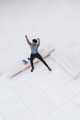 Happy man in casual wear overjoyed with finish of startup project developing with modern laptop computer, accomplished Latino male freelancer with raised hands celebrating approved remote work