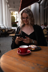 A stylish girl of twenty-five years old sits in a cafe on the outdoor terrace, drink coffee and eat croissant, the woman is wearing a black dress, jacket and glasses,