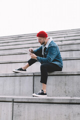 Trendy dressed hipster guy typing text message or content publication for social networks while sitting at city stairs, talented man with stylus drawing sketch during distance job outdoors
