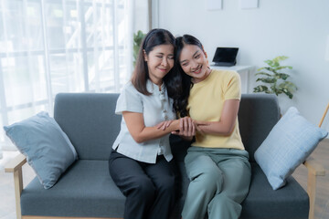 Obraz premium Asian mother and daughter are holding hands and smiling, sitting on a sofa in the living room, enjoying a tender moment of affection and complicity