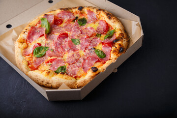 Pizza with cheese, ham, green basil leaves and tomatoes in paper box on black background. Food product advertising banner, copy space