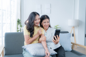 Cheerful senior mother and adult daughter making a video call with smartphone sitting on sofa in the living room at home, they're smiling and waving