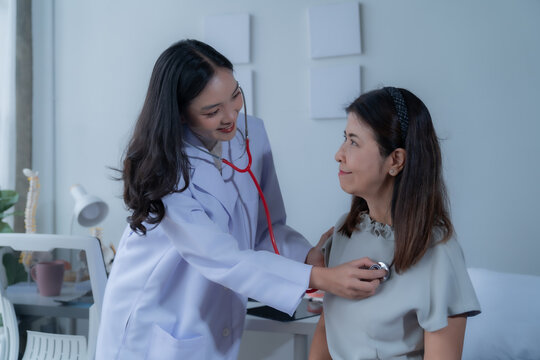 Smiling female doctor using a stethoscope to examine the lungs and heart of a middle-aged female patient during a medical checkup in a bright, welcoming clinic environment