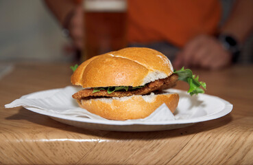 Crispy chicken sandwich on a white plate, lettuce garnish, man holding beer in background, fast food concept