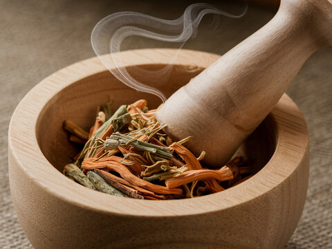 A wooden mortar and pestle is used to crush herbs, releasing aromatic scents into air. gentle smoke adds calming atmosphere to scene, enhancing experience of preparation