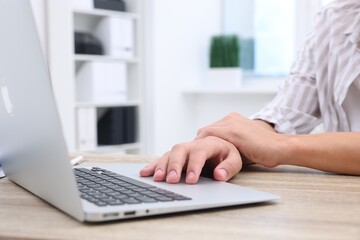 Man suffering from pain in wrist while working on laptop at table indoors, closeup. Carpal tunnel...