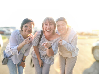Three friends stand close together with bright smiles, enjoying a sunny evening outdoors in a lively parking area