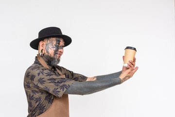 Side view of a unique looking barista handing over takeaway coffee. Wearing a black hat, full of face tattoos and piercings. Isolated on a white background.