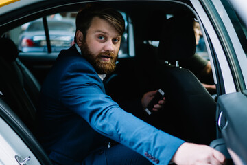 Portrait of handsome bearded man in formal suit looking at camera while getting to business meeting in financial district of Manhattan, Caucasian corporate director sitting on passenger seat in taxi