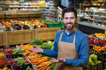 Smiling grocer wearing apron presenting fresh organic vegetables in a grocery store. Surrounded by colorful produce including carrots, radishes, and greens, promoting healthy food choices.