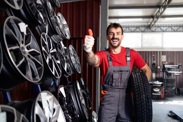 Smiling worker holding new tire and standing inside wheel repair shop.