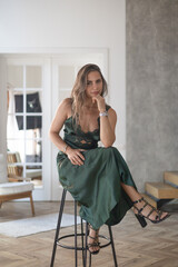 Beautiful fashionable middle aged woman in elegant green silk dress sitting in armchair at home