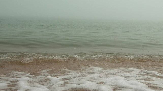 Mother-of-pearl sea surf. A quiet foggy sea, sandy-shell beach, the sun breaks through the fog and colors the crest of the rolling waves