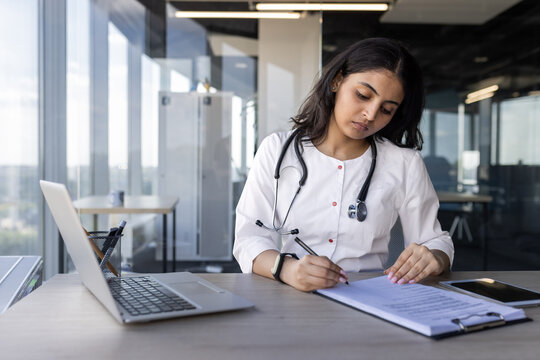 Serious thinking female doctor behind paperwork inside office, female clinic worker records patient data, enters data into forms and documents, uses laptop at work, prepares report