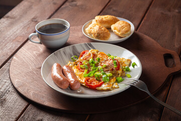 Eggs with tomatoes and sausages in a plate on a wooden table of coffee and croissants on a round stand