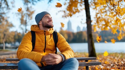 A young man in a beanie and jacket, surrounded by falling autumn leaves, sitting on a park bench, embodying joy and the beauty of nature during fall.
