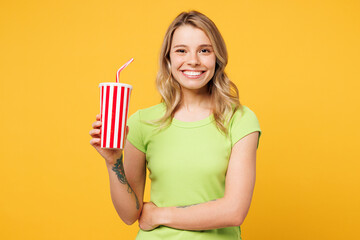 Young happy blonde woman wear green t-shirt casual clothes hold in hand cup of soda pop cola fizzy water looking camera isolated on plain yellow orange background studio portrait. Lifestyle concept.