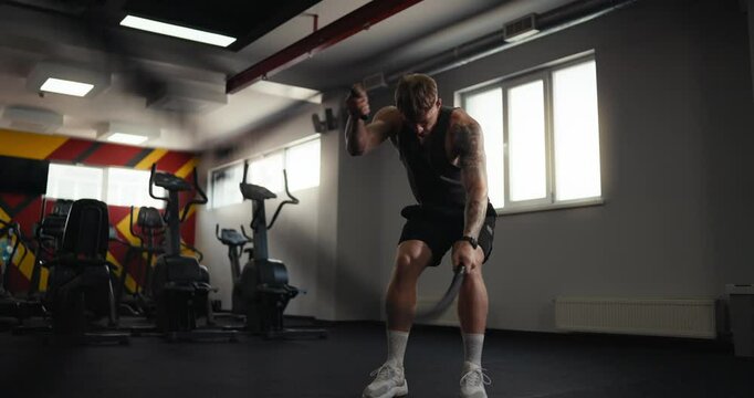Confident sporty blond guy with tattoos in black sportswear performs exercises with two ropes to strengthen arm muscles in the gym