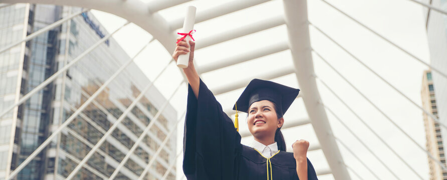 Graduate and Success Education in University Concept. Happy student woman graduate diploma degree in asian college.Young graduate in cap and gown raising diploma against modern cityscape background.