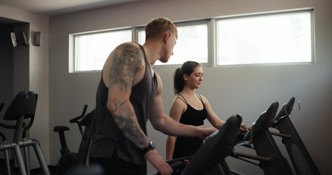 Confident guy trainer athlete with tattoos in a gray T-shirt tells a female client how to properly set up a treadmill for a comfortable workout in the gym