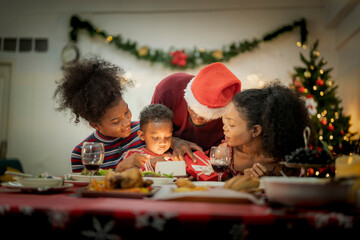 A joyful family gathers around the dinner table celebrating Christmas. The father, dressed in a Santa hat, gives a gift to the child, while everyone smiles with warmth and festive decorations.