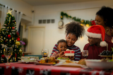 A joyful family gathers around the dinner table celebrating Christmas. The father, dressed in a Santa hat, gives a gift to the child, while everyone smiles with warmth and festive decorations.