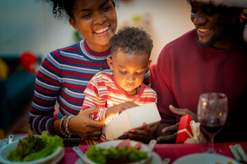 A heartwarming Christmas moment as a young child opens a beautifully wrapped gift with the help of his parents. The mother gently guides the child while the father, dressed in a Santa hat.