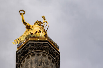 A statue of a woman holding a wreath on top of a building. The statue is gold and the sky is cloudy
