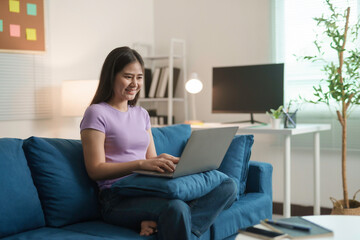 Naklejka premium Young woman sits on a blue sofa in her living room, happily working on her laptop