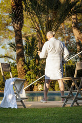 A man of retirement age in a white robe by the pool against an exotic landscape with palm trees and a pool enjoying the setting sun.
