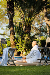 A man of retirement age in a white robe by the pool against an exotic landscape with palm trees and a pool enjoying the setting sun.

