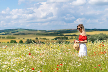 Blonde young woman in sunglasses, red and white dress in a wildflower field, holding a straw hat.