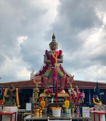 Giant Thai Guardian Statue at Temple Shrine with Colorful Offerings and Cloudy Sky Background